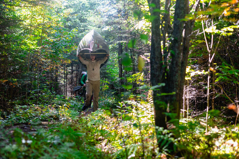 A person is carrying a canoe over their head while walking through a lush green forest. The individual is wearing light-colored pants and a long-sleeved shirt. Sunlight filters through the trees, creating dappled light on the forest floor. The canoe appears to be green or camouflaged, blending with the surrounding foliage. The scene suggests an outdoor adventure or portage during a canoeing trip.